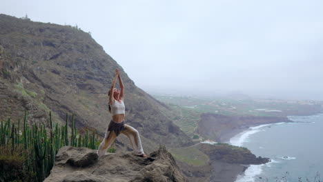 Against-the-backdrop-of-the-blue-ocean-at-sunset,-a-young-woman-performs-yoga-on-a-rocky-seashore,-embodying-a-healthy-lifestyle,-harmony,-and-the-interplay-between-humans-and-nature